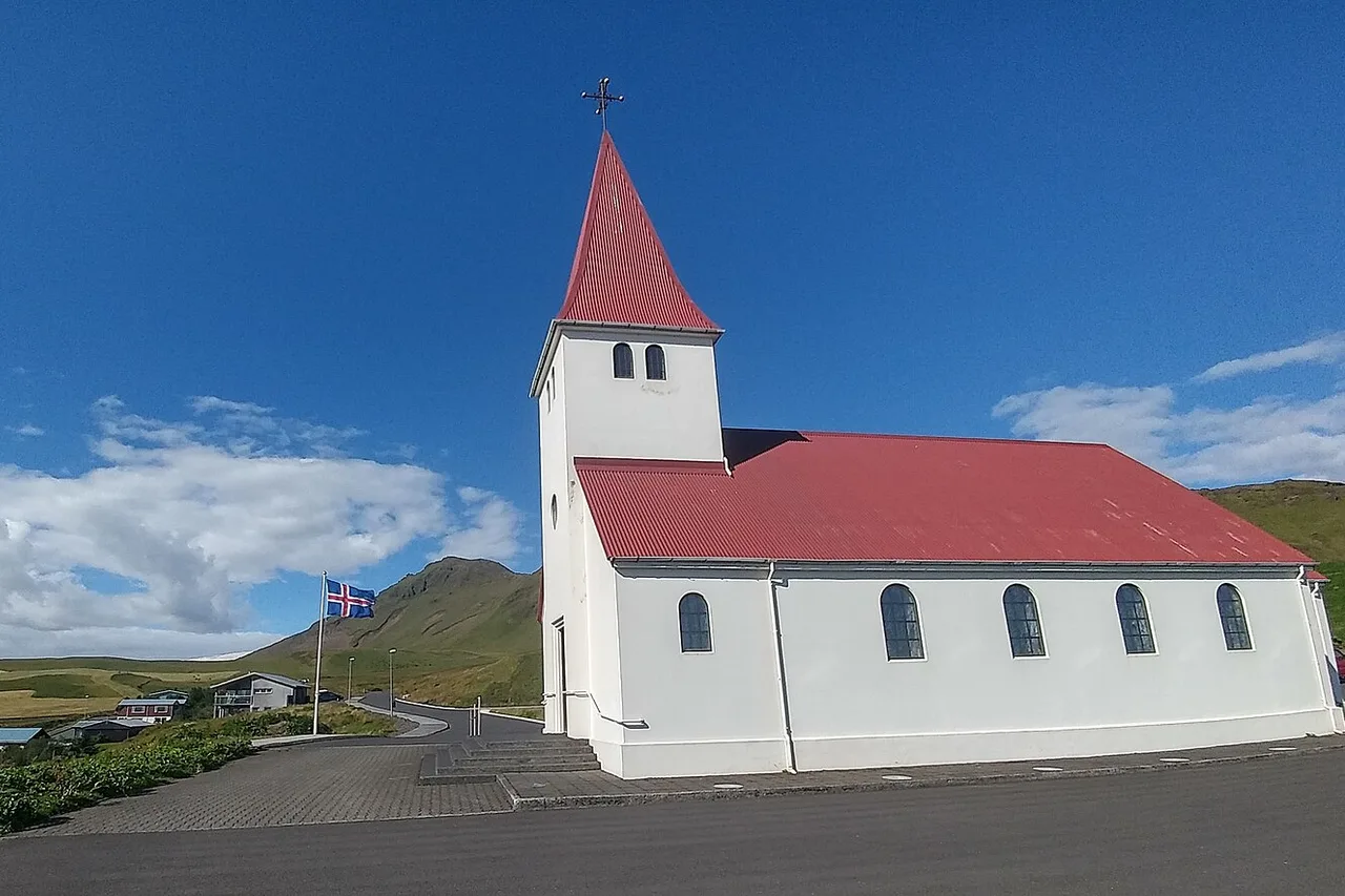L’église de Vík í Mýrdal : un édifice blanc au toit rouge et au clocher, le drapeau islandais devant, des collines vertes derrière sous un ciel bleu.