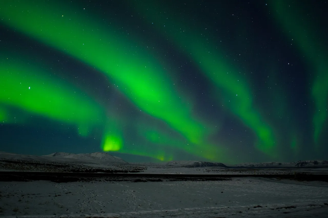 Grüne Polarlichter wölben sich in Bögen über einer verschneiten Ebene und fernen Bergen in Island.