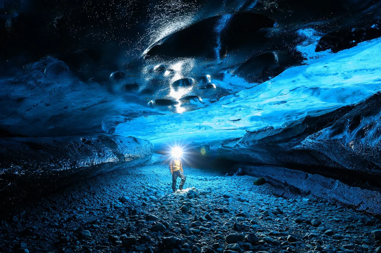 Ein Mensch mit Stirnlampe steht in einer blau leuchtenden Eishöhle, über ihm wölbt sich die glatte Eisdecke.