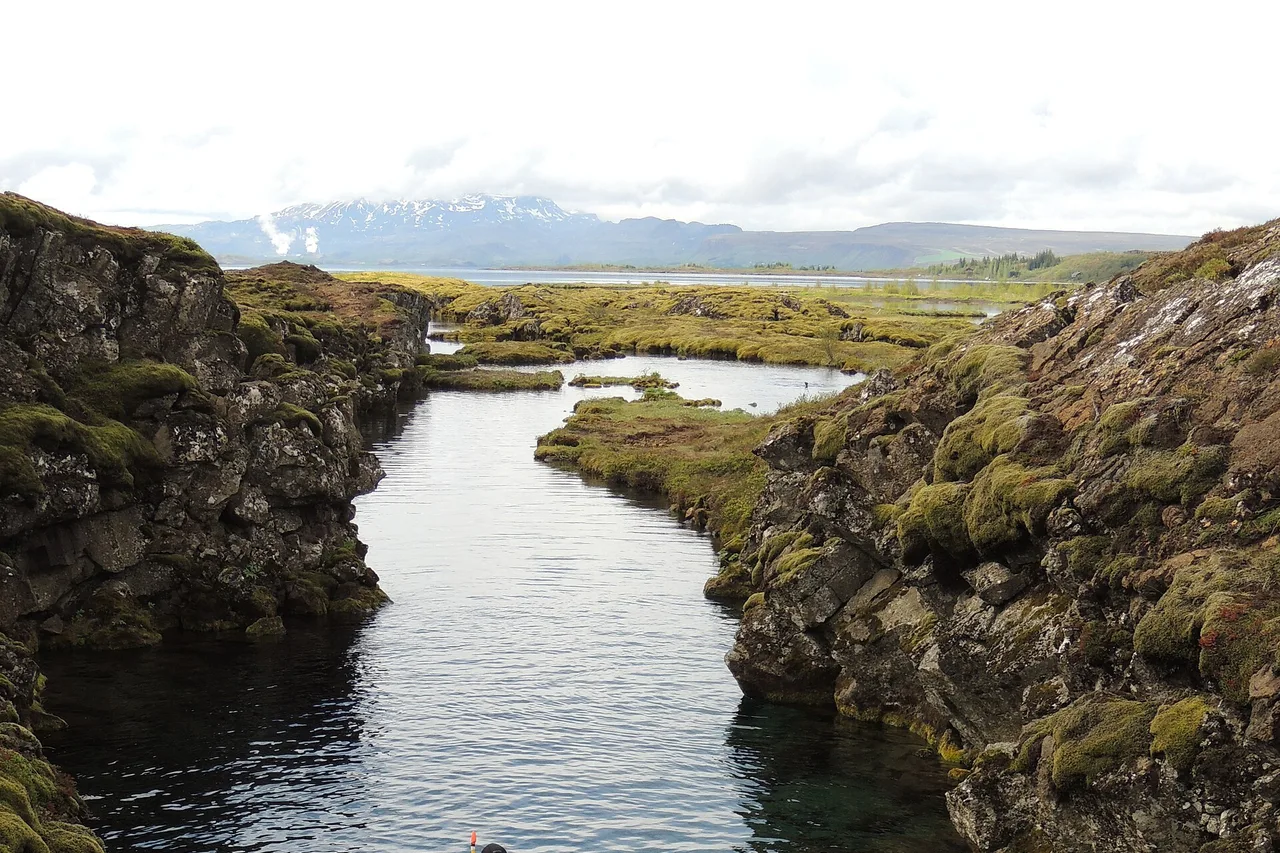 Die schmale Silfra-Spalte zwischen bemoosten Lavawänden, dahinter der See und ferne Berge; am unteren Bildrand ragt der Schnorchel eines Schwimmers aus dem Wasser.