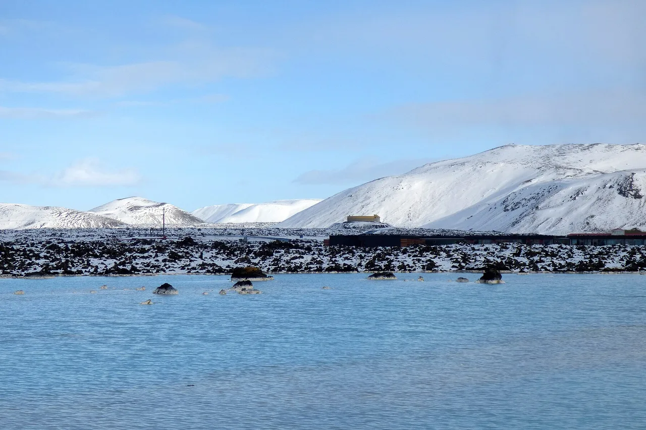 Eau bleu pâle du Blue Lagoon bordée de lave noire couverte de neige, montagnes blanches au fond