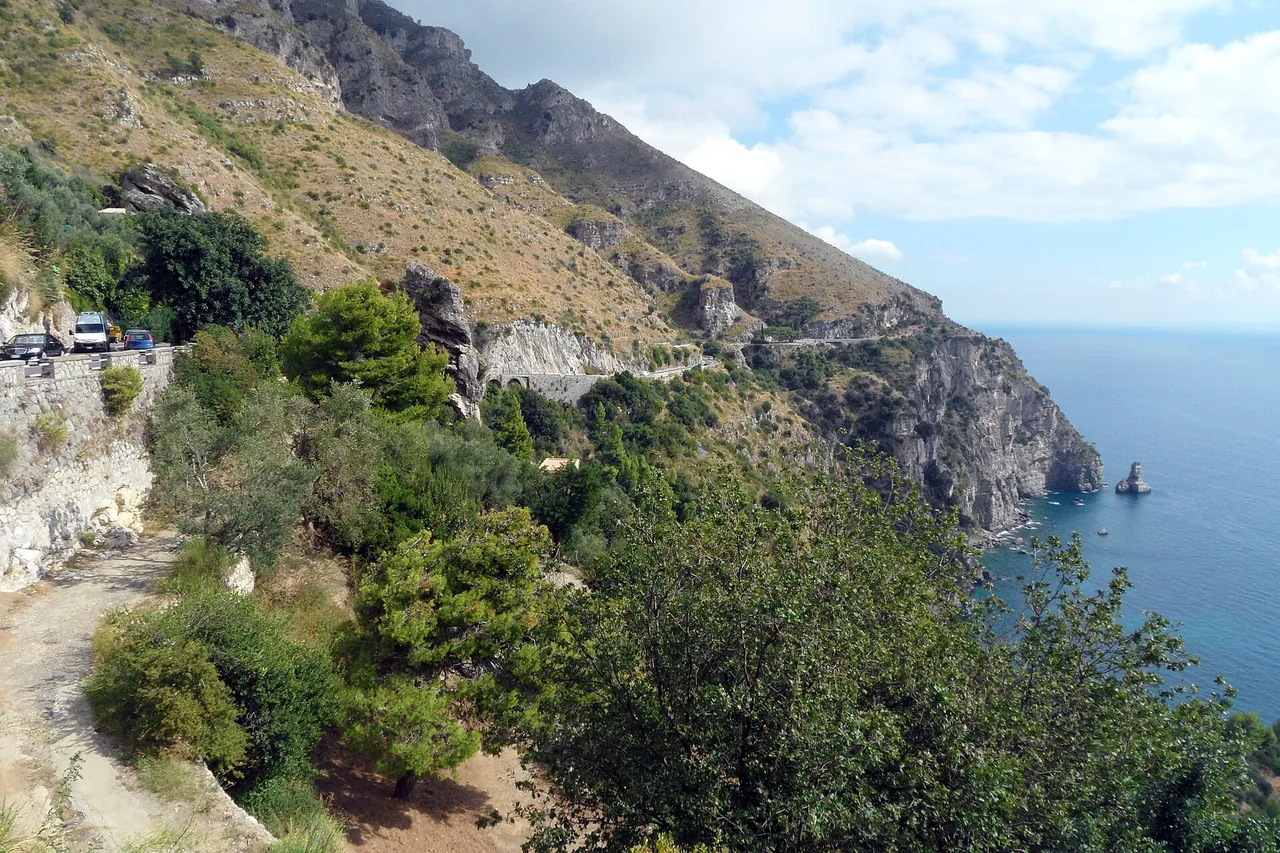 Une route côtière taillée dans la pente sur la côte amalfitaine, avec un minibus et des voitures à gauche et la mer bleue à droite, un rocher dans l’eau