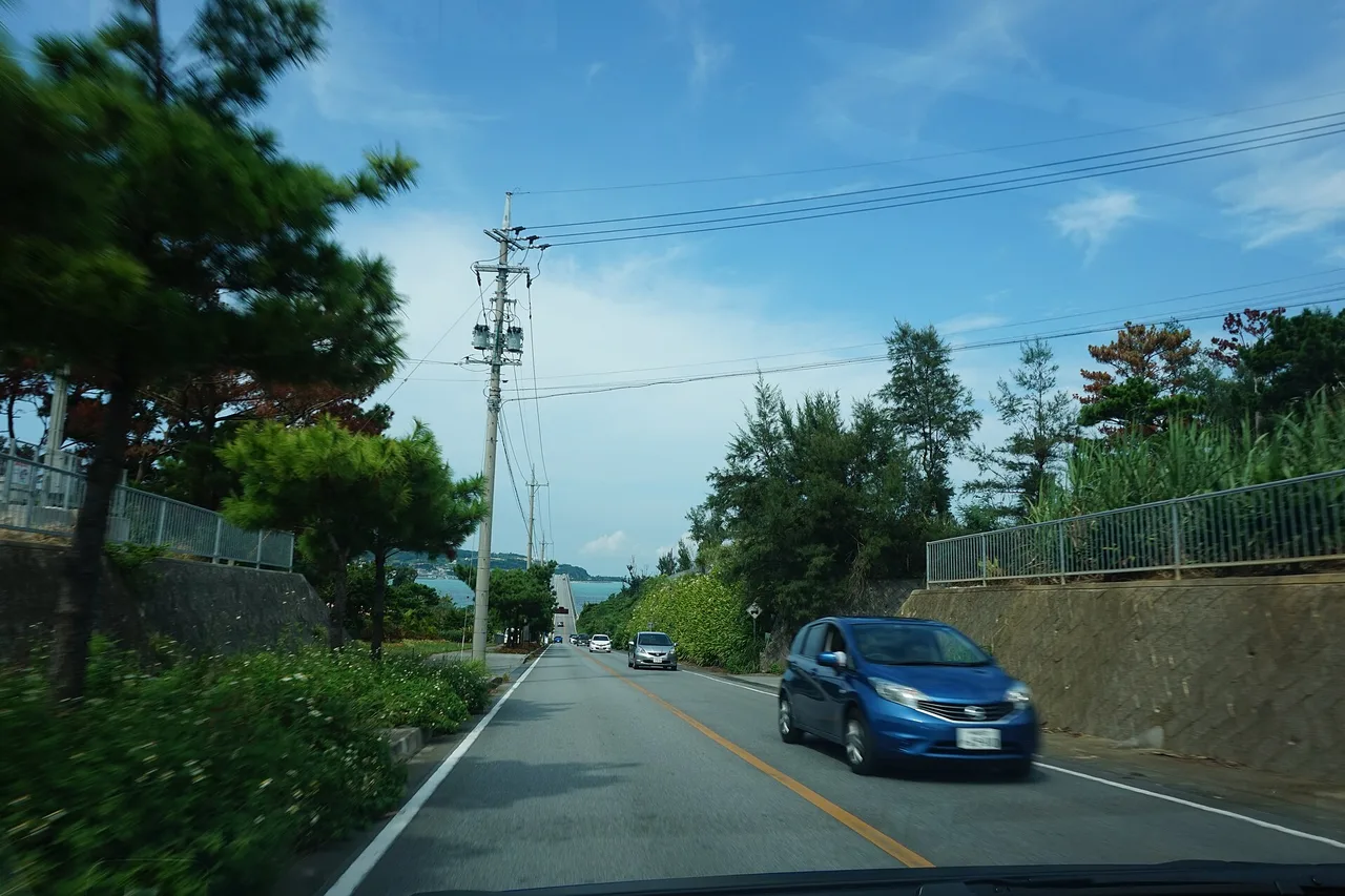 Una strada prima del ponte di Kouri a Okinawa, con guida a sinistra: una piccola auto blu viene incontro, sullo sfondo il mare