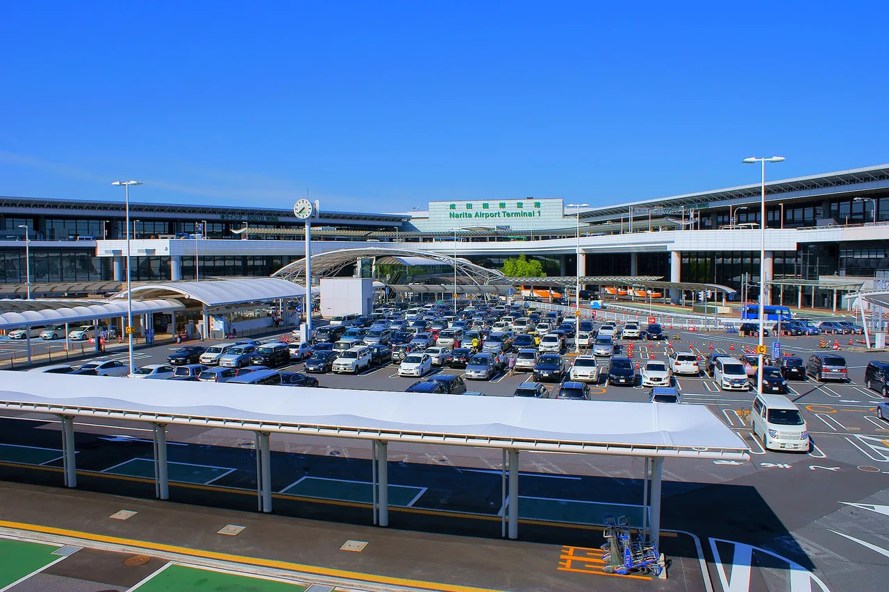 Terminal 1 des Flughafens Narita mit Uhr, Schriftzug über dem Eingang und einem vollen Parkplatz davor
