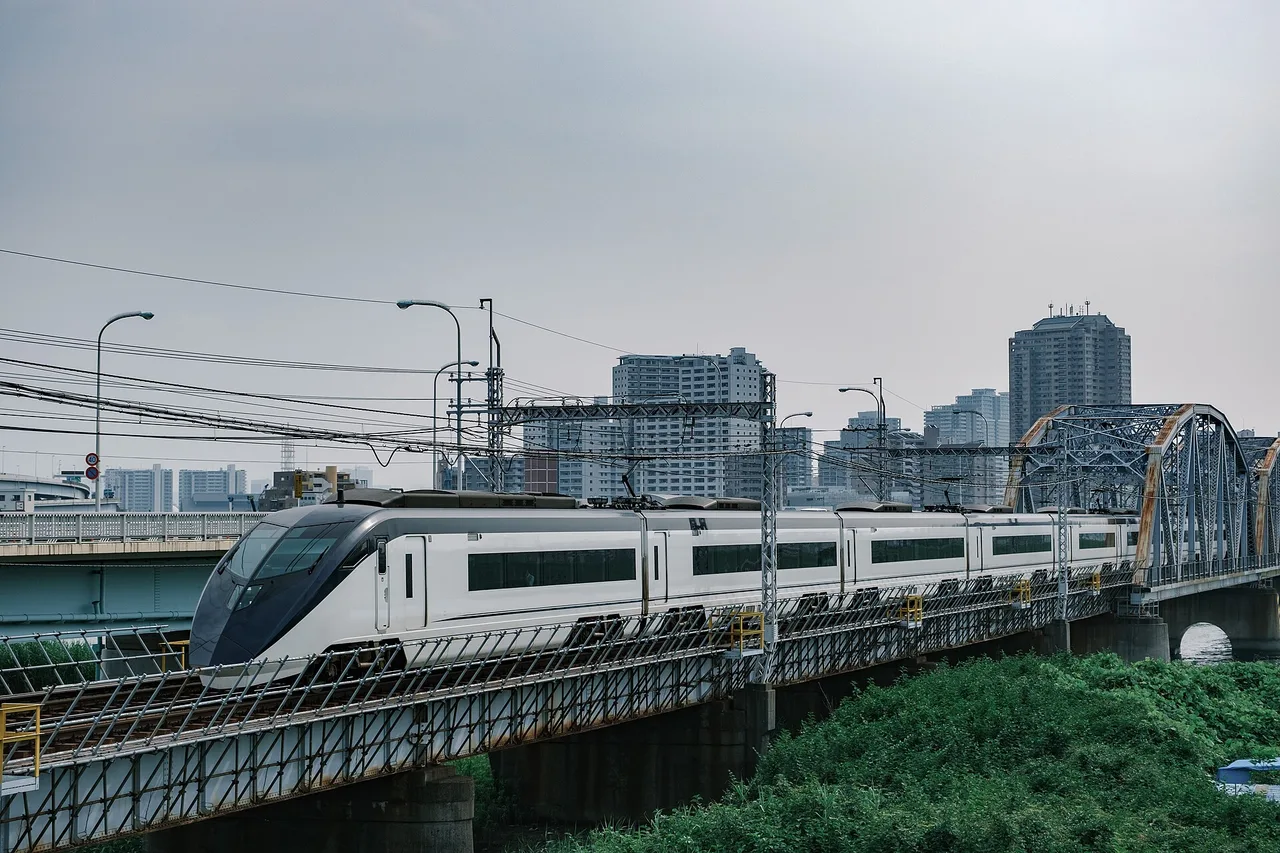Ein weißer Skyliner-Zug von Keisei auf einer Eisenbahnbrücke über einem Fluss, im Hintergrund Wohnhochhäuser