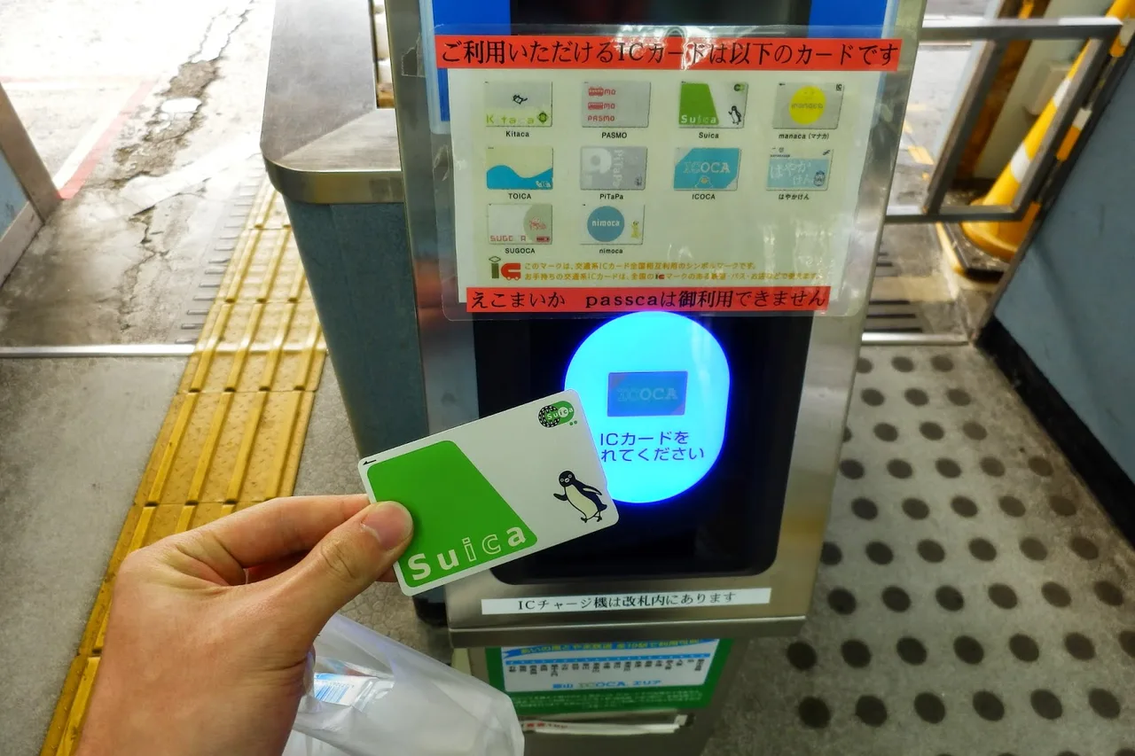 A hand touching a green Suica card onto the blue reader of a station ticket gate