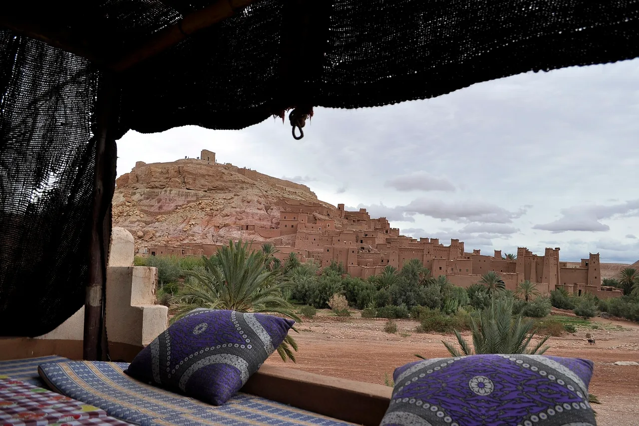 The ksar of Ait Benhaddou seen from a terrace shaded by a reed canopy, with cushions in the foreground