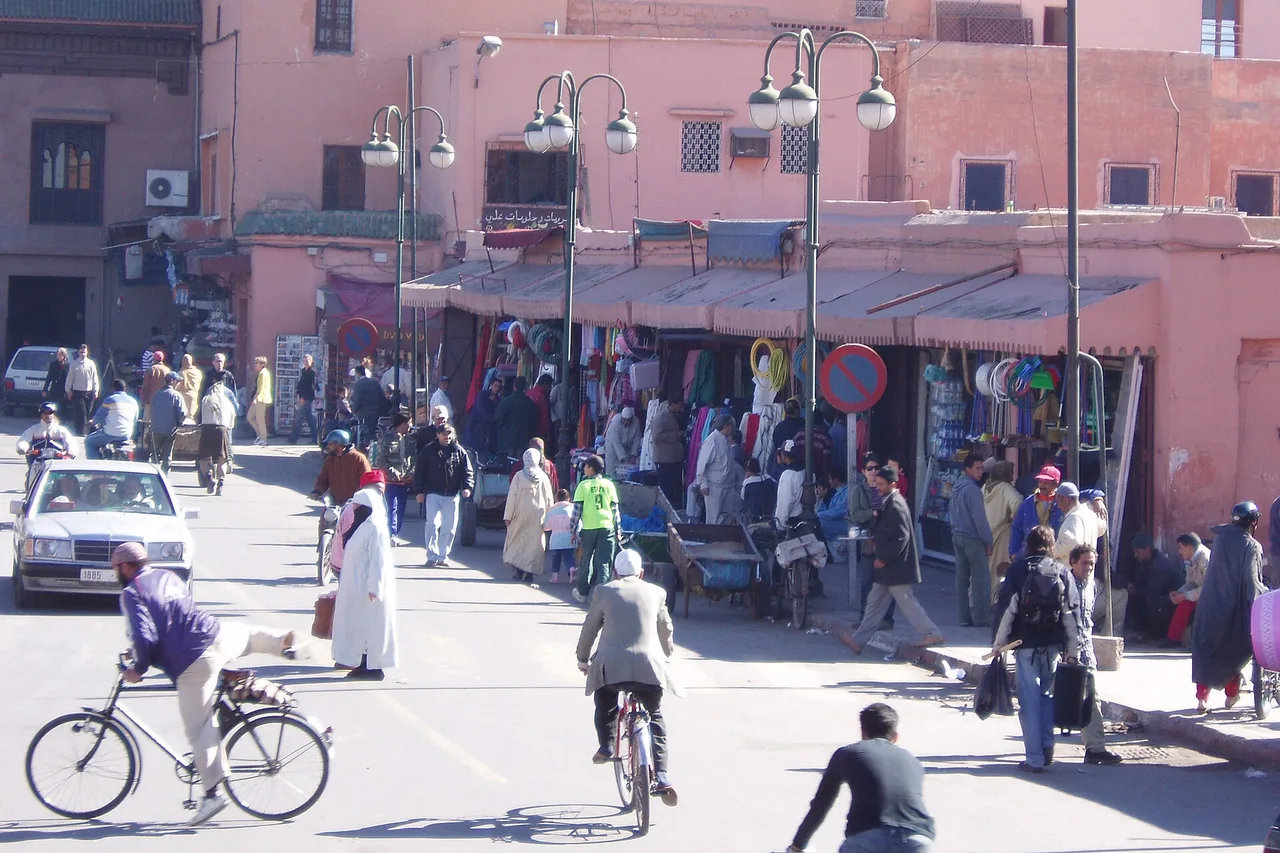 A wide street in the Marrakech medina with pink-ochre walls, cyclists and pedestrians in full sun