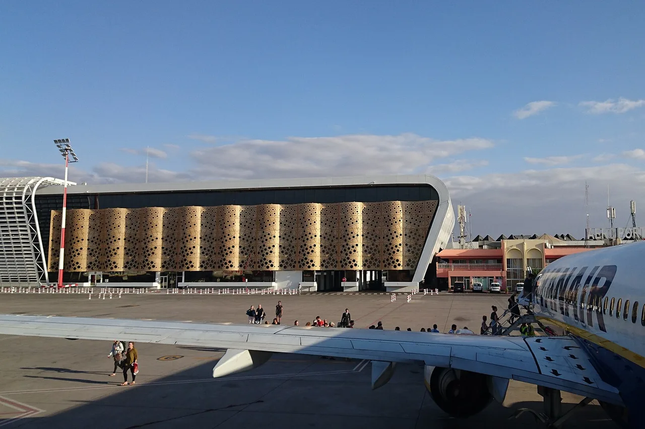 Terminal del aeropuerto de Marrakech-Menara con fachada dorada calada, en primer plano el ala de un avión y pasajeros en la pista