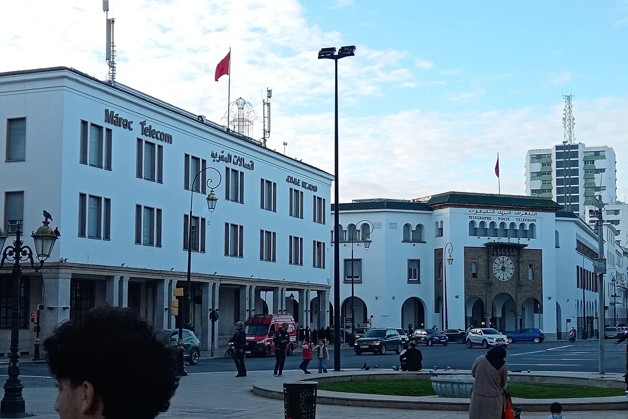 Edificio blanco con el rótulo Maroc Telecom en una plaza, a la derecha el edificio de correos con un reloj, peatones y coches delante