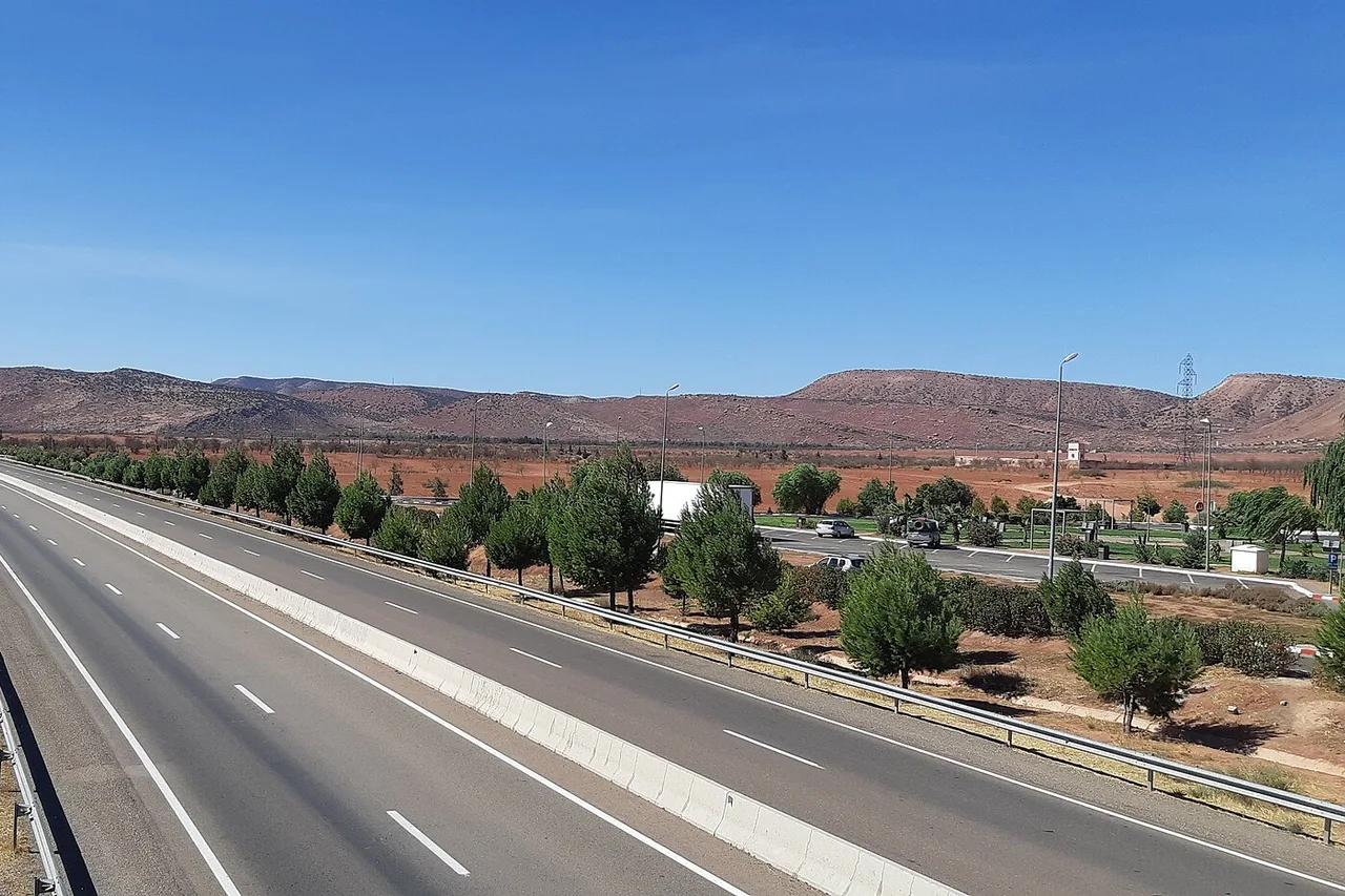 Una autopista vacía de calzadas separadas atraviesa unos árboles hacia colinas rojizas, la autopista entre Marrakech y Agadir