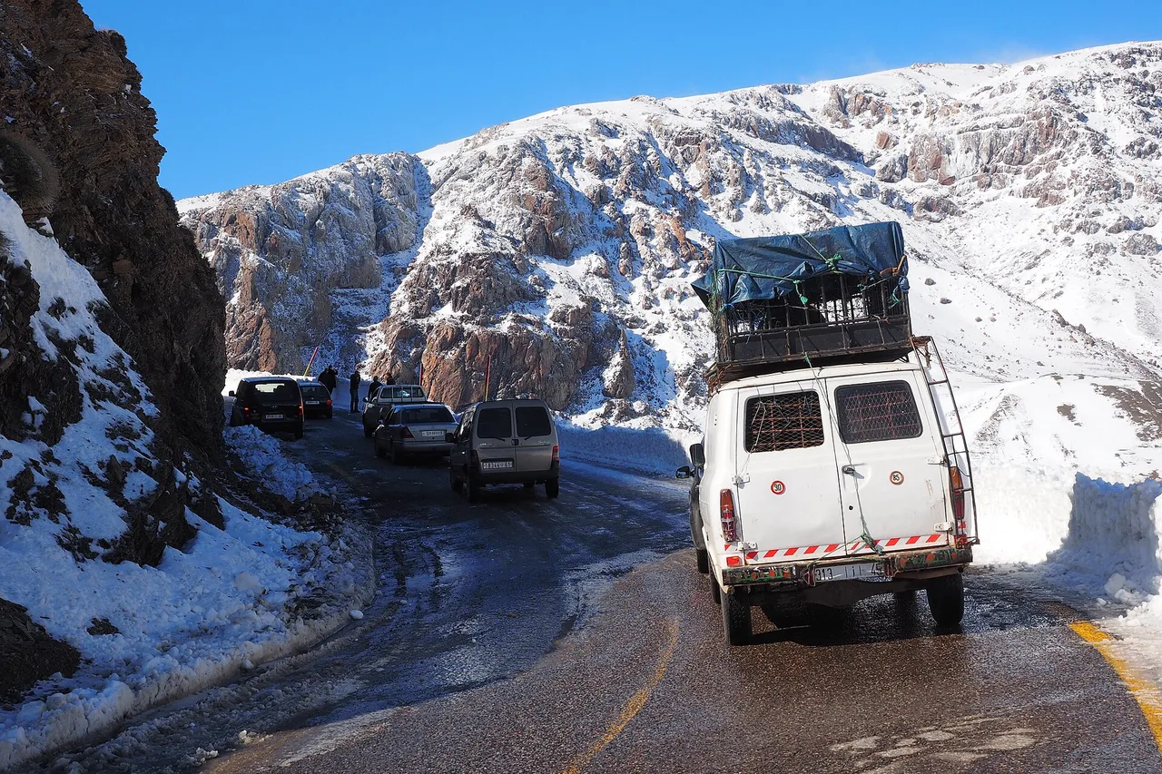 Una furgoneta blanca con equipaje en el techo circula por una carretera de montaña entre montones de nieve, con otros vehículos delante de paredes rocosas nevadas en el Alto Atlas