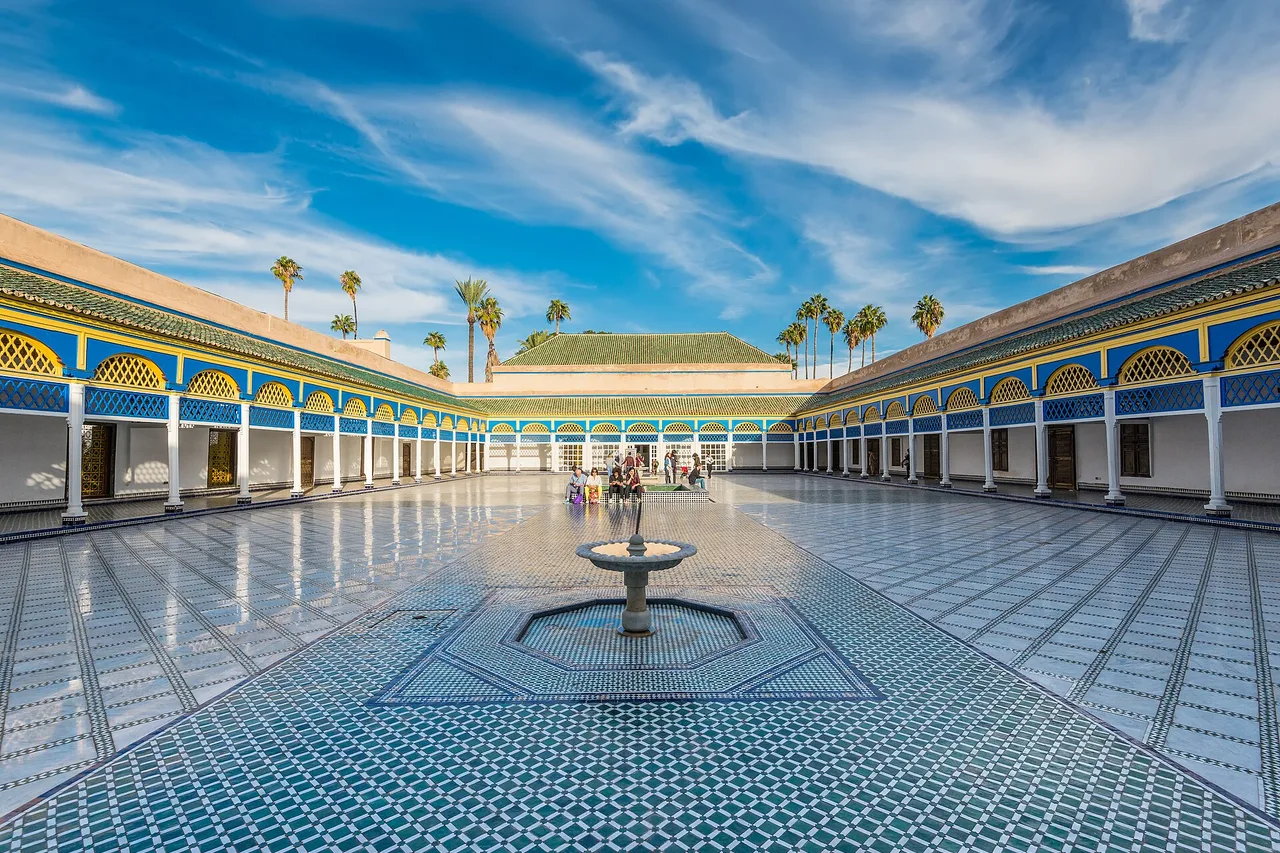 La grande cour dallée du palais de la Bahia, avec une fontaine au centre, des arcades peintes aux motifs bleus et des palmiers sous un ciel bleu