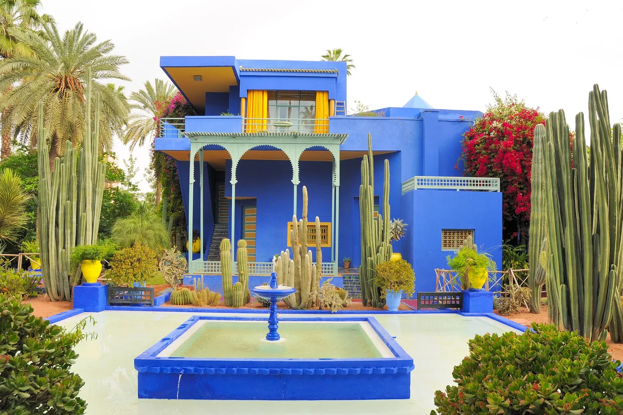 La villa bleu cobalt du jardin Majorelle à Marrakech, avec ses volets jaunes, un bassin bleu au premier plan, de grands cactus et des palmiers