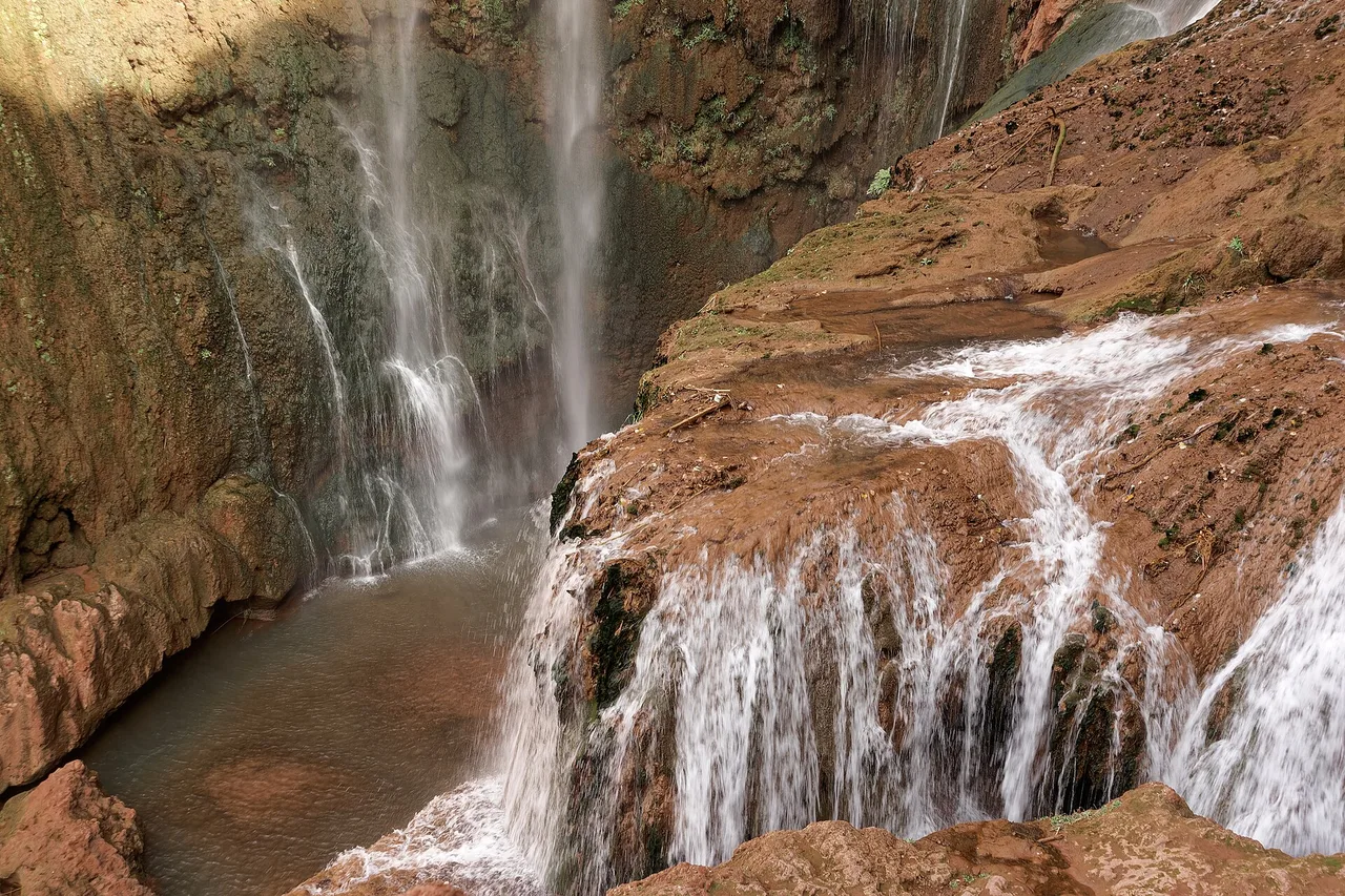 Une cascade tombe sur une roche argileuse rouge dans les chutes d’Ouzoud, avec quelques visiteurs au bord d’un bassin sur la gauche