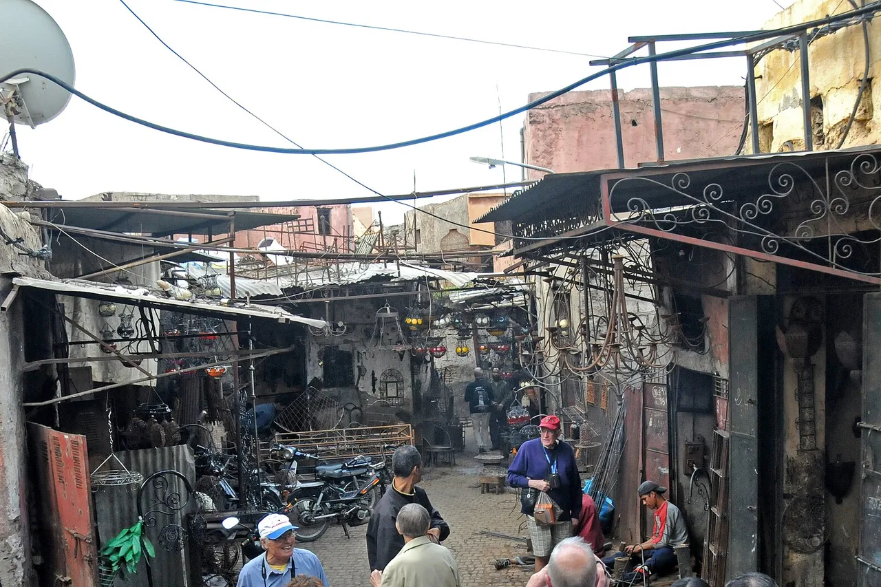 Une ruelle étroite de la médina de Marrakech avec des ateliers et des échoppes, des câbles au-dessus des têtes, des mobylettes garées et quelques passants