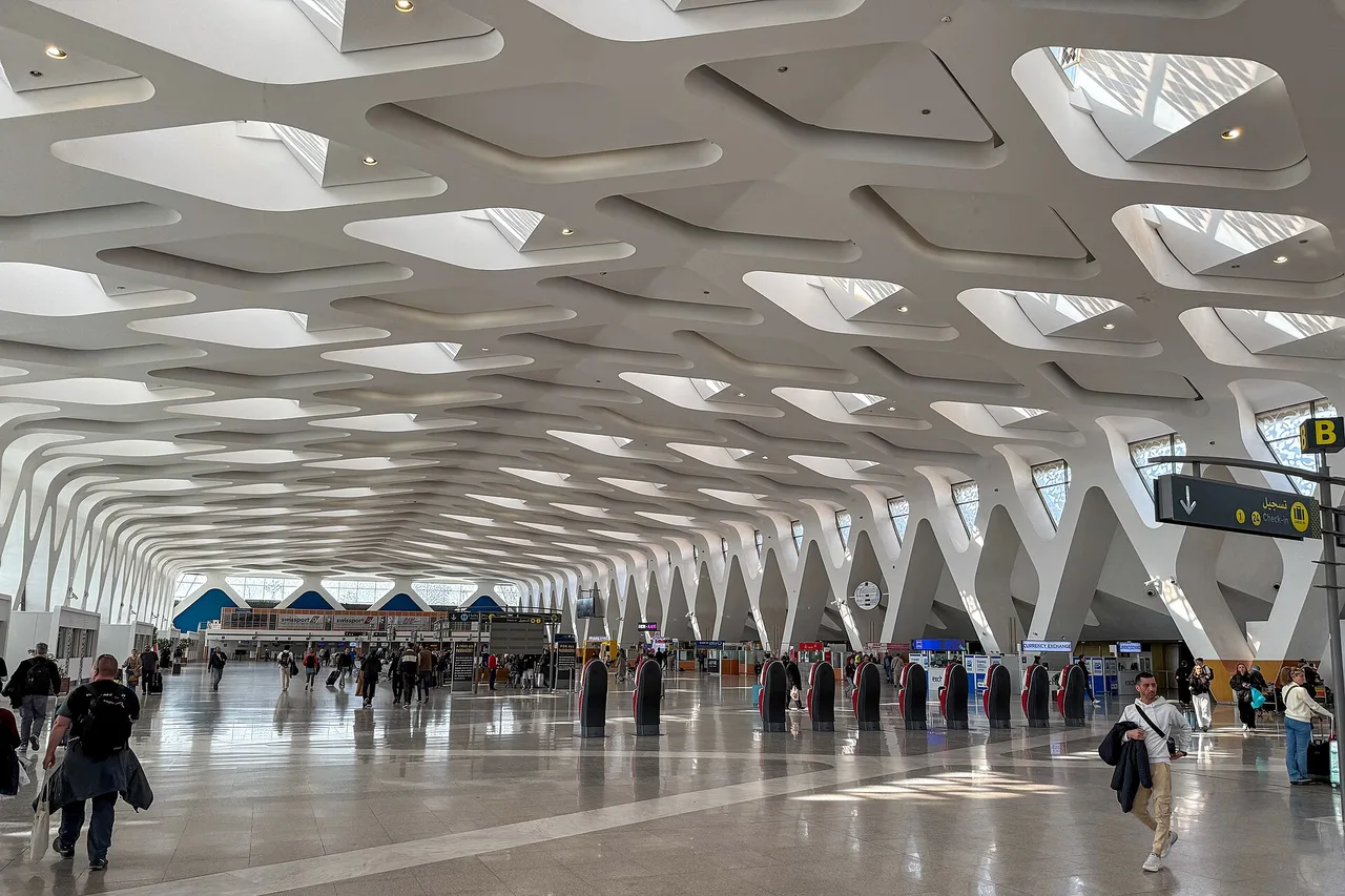 Grande sala del terminal dell’aeroporto di Marrakech-Menara con soffitto bianco a struttura traforata, viaggiatori con i bagagli e banchi sullo sfondo