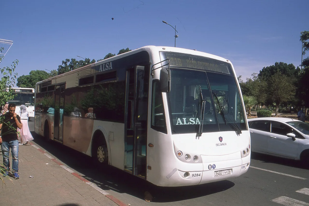 Bus ALSA bianco fermo lungo un marciapiede a Marrakech, passanti e alberi sullo sfondo