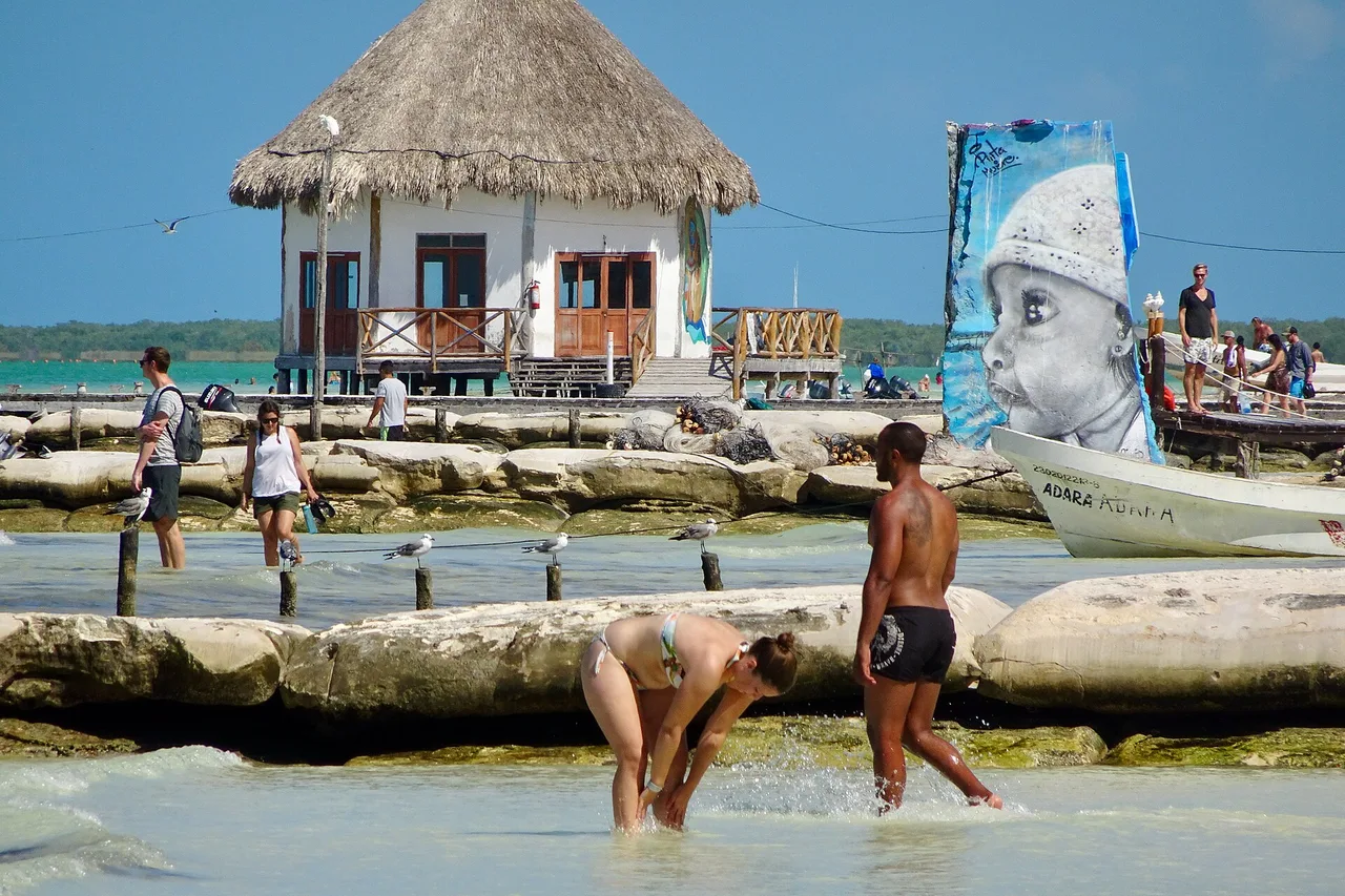 Strand von Holbox mit Holzsteg, strohgedecktem Haus auf Stelzen, Booten und Badegästen im flachen Wasser