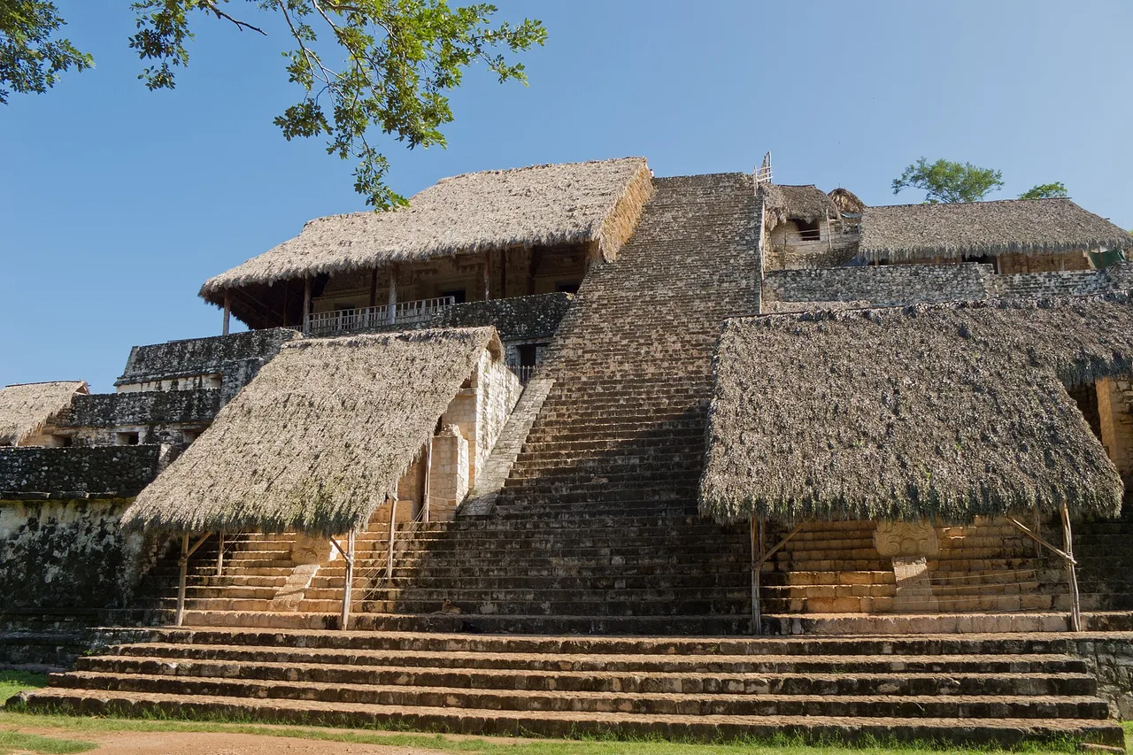 The Acropolis of Ek Balam: a wide stepped pyramid topped with palm roofs protecting the stucco, lawn in the foreground.