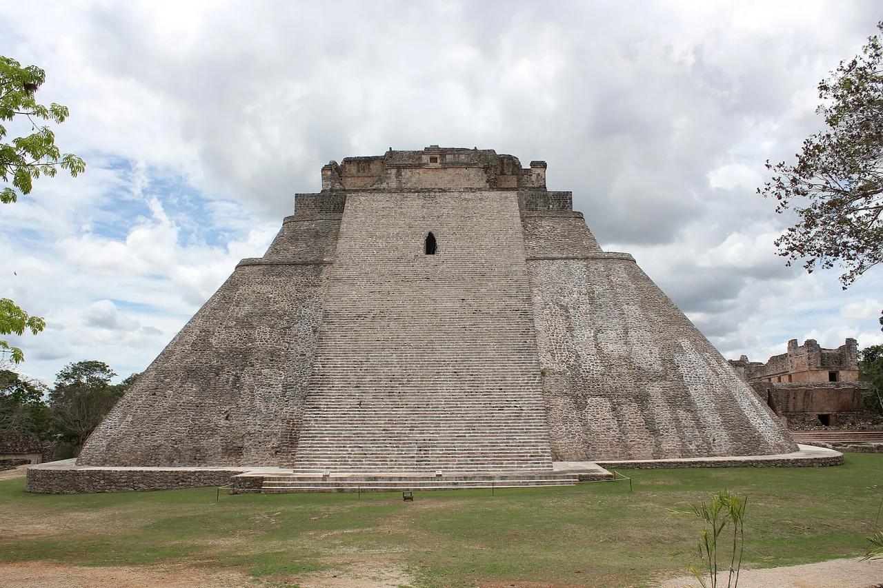 The Pyramid of the Magician at Uxmal: an oval stepped building of pale limestone with a steep stairway under a blue sky with clouds.