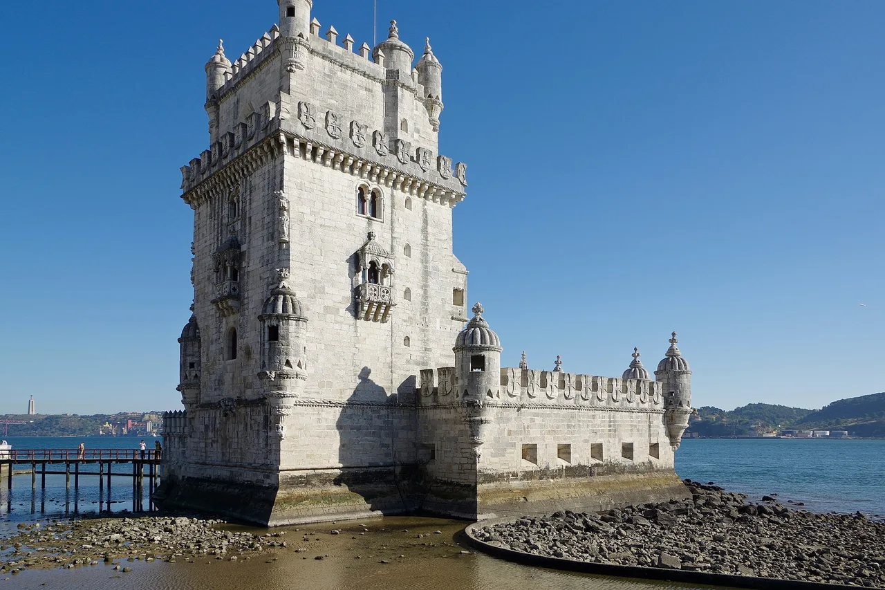 Belém Tower on the bank of the Tagus, a pale crenellated tower with small corner turrets, rocks and water in front of it under a blue sky