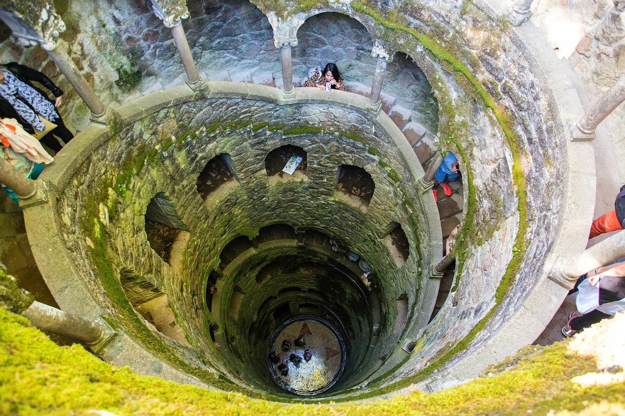 Looking down into the Initiation Well at Quinta da Regaleira, a stone spiral staircase with moss-covered walls winding down into the depths