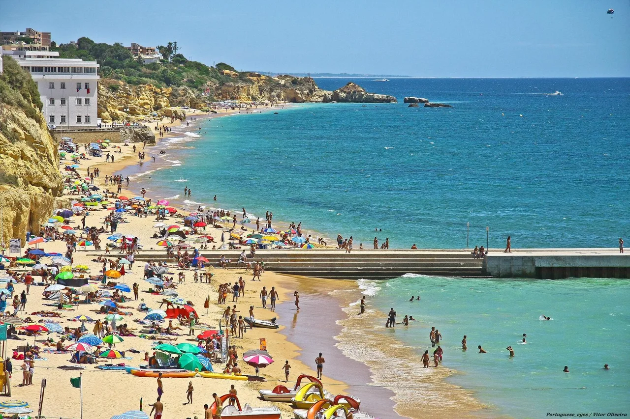 Praia dos Pescadores in Albufeira an einem Sommertag, mit Sonnenschirmen, Badegästen und einer Mole im türkisfarbenen Wasser