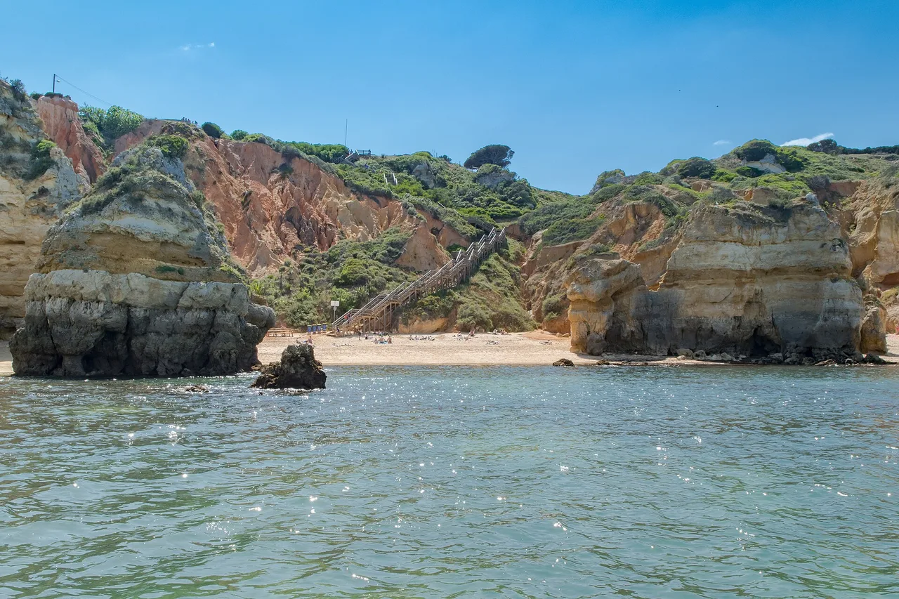 Praia do Camilo seen from the sea, between two limestone stacks, with the long wooden staircase running down the cliff