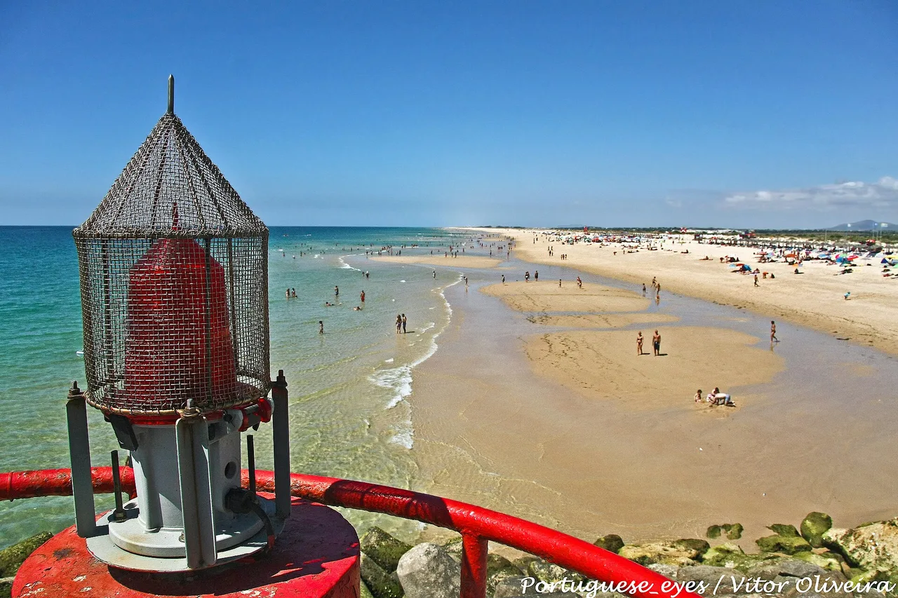 A red lighthouse in the foreground and the wide sandbank of Tavira island beach behind it, in shallow water
