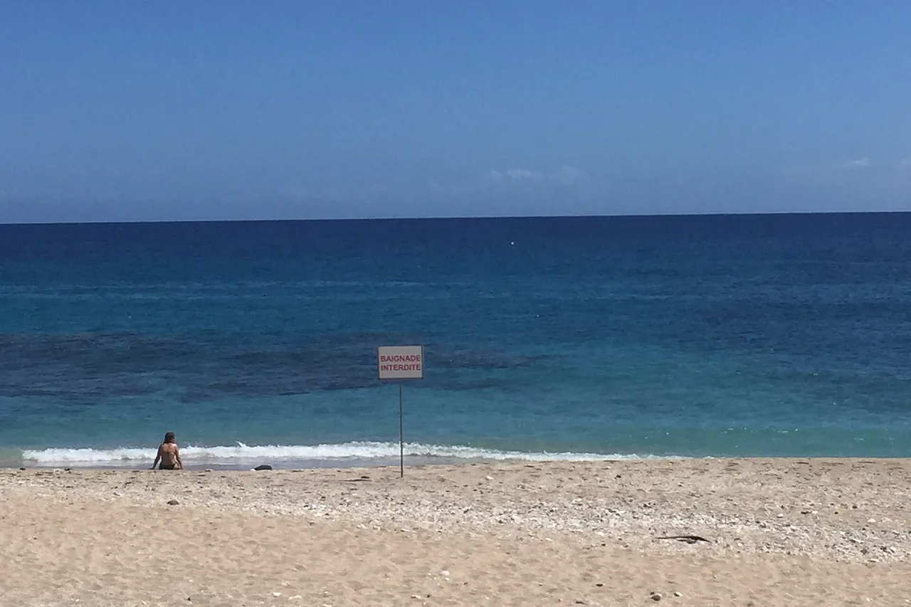 Badeverbotsschild im Sand von Boucan Canot, türkisfarbenes Wasser im Hintergrund, La Réunion