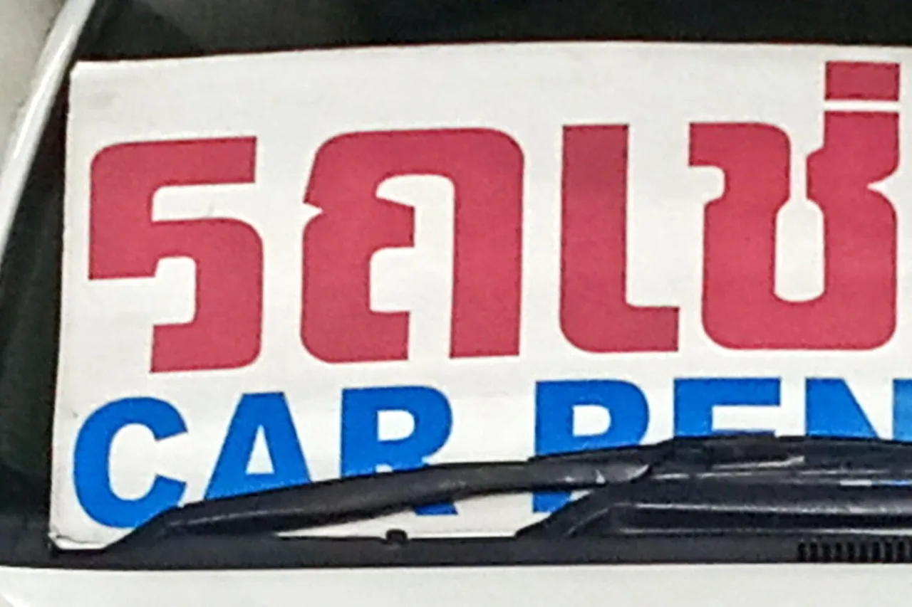 Ein zweisprachiges Schild mit der Aufschrift „car rent“ in Thai und Englisch hinter einer Windschutzscheibe