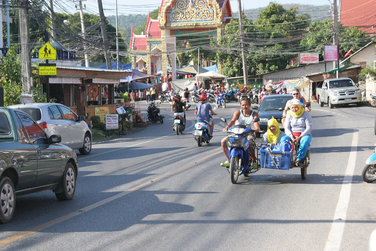 Eine Straße auf Phuket mit Autos, Rollern und einem Motorrad mit Beiwagen, im Hintergrund ein Tempeltor