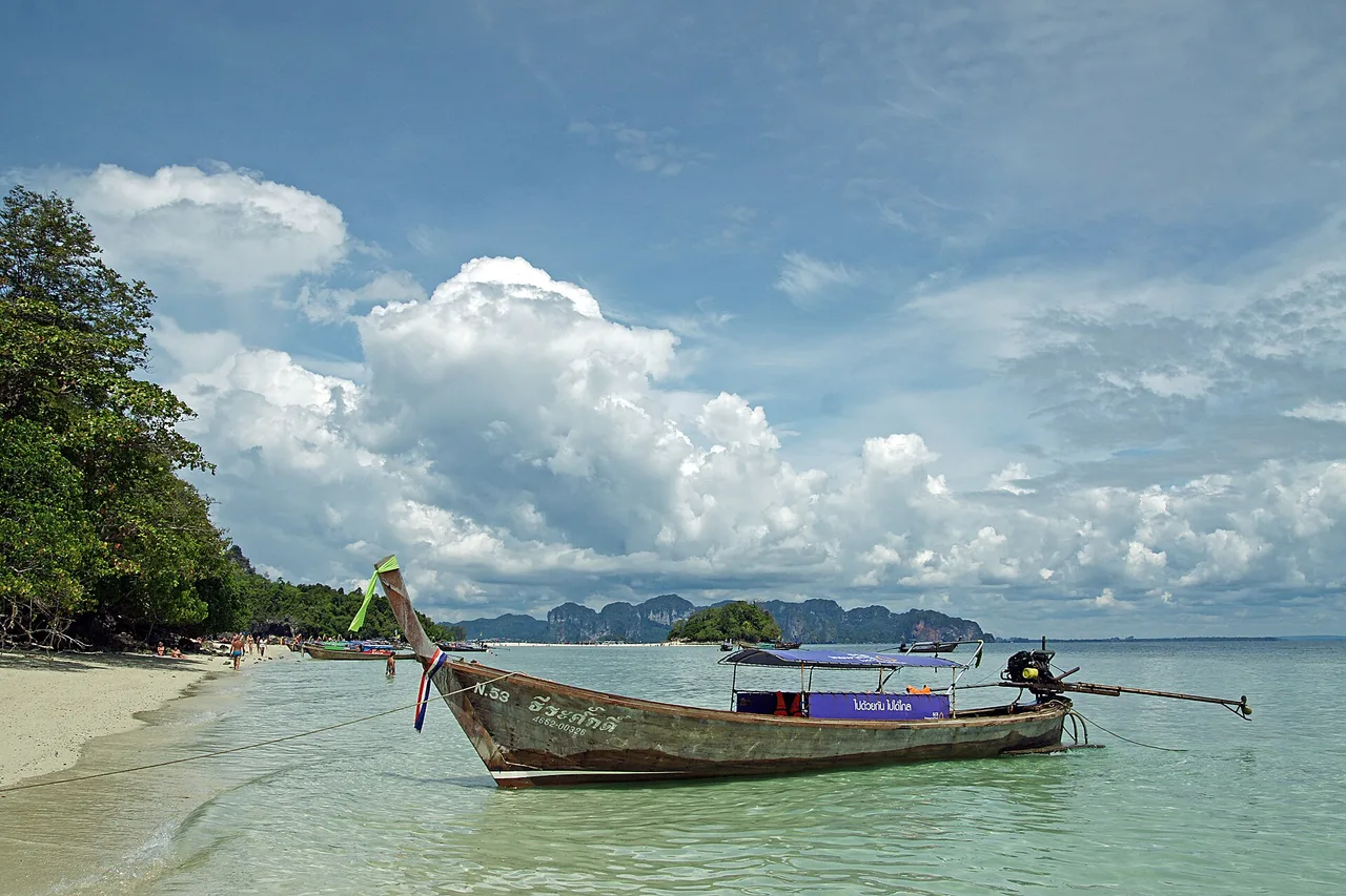 Un bateau à longue queue échoué sur la plage de sable de Ko Kai, près de Krabi, avec une mer plate, des îles lointaines et de gros cumulus derrière