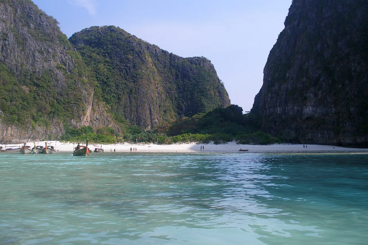 Une plage de sable clair à Maya Bay au pied de hautes parois calcaires couvertes de végétation, avec quelques bateaux à longue queue près du rivage dans une eau turquoise