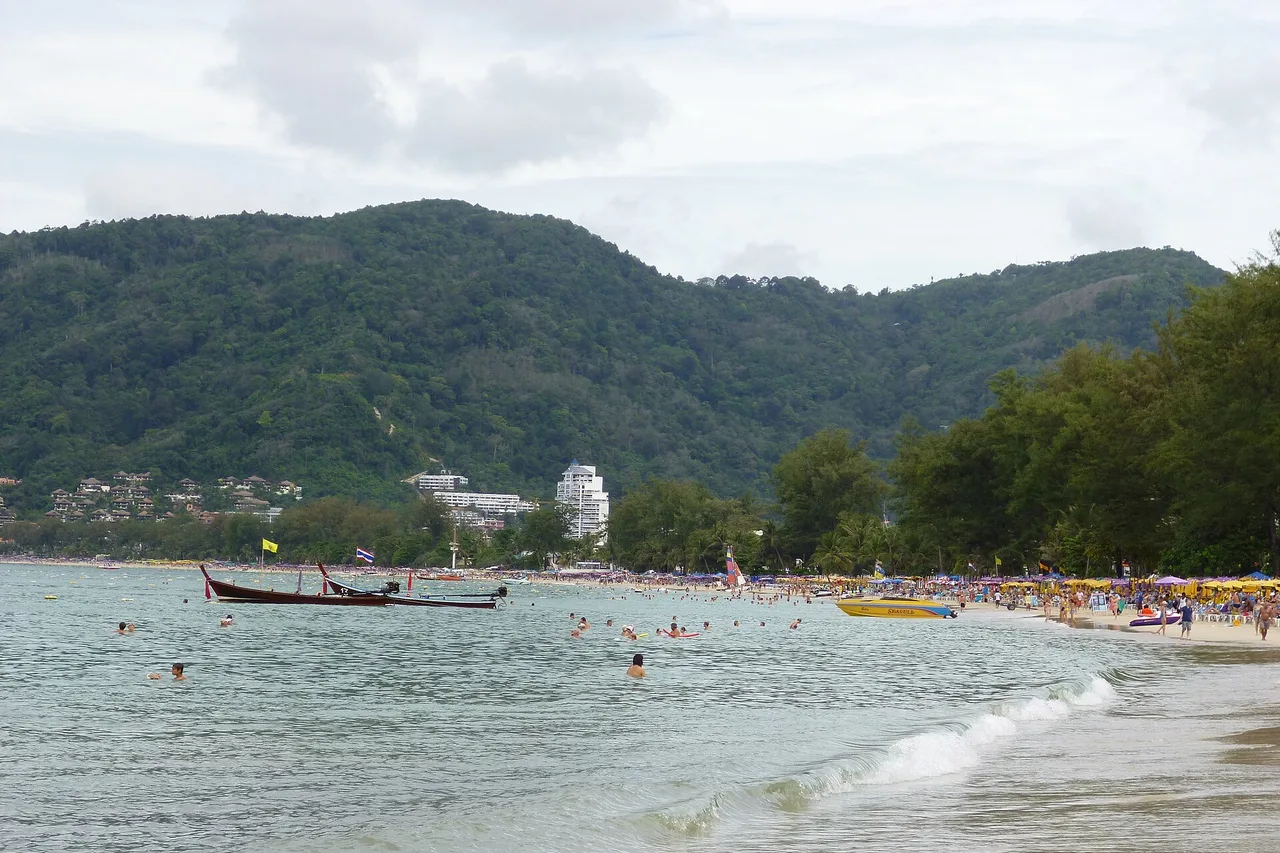 Die Bucht von Patong mit Langschwanzboot im grauen Wasser, Sonnenschirmen am Strand und bewaldeten Hügeln dahinter