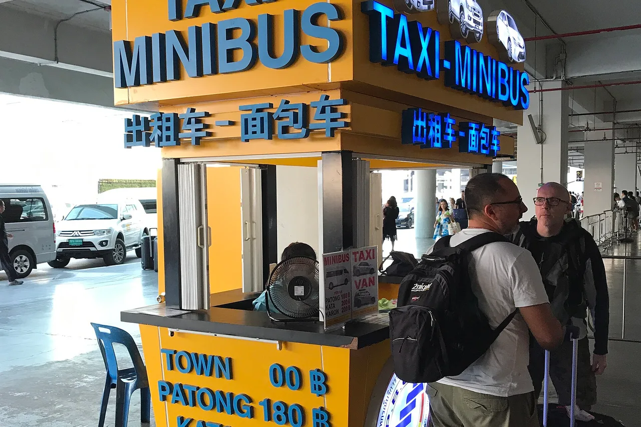 Gelber Taxi-Minibus-Schalter in der Halle des Flughafens Phuket, davor zwei Reisende mit Rucksack