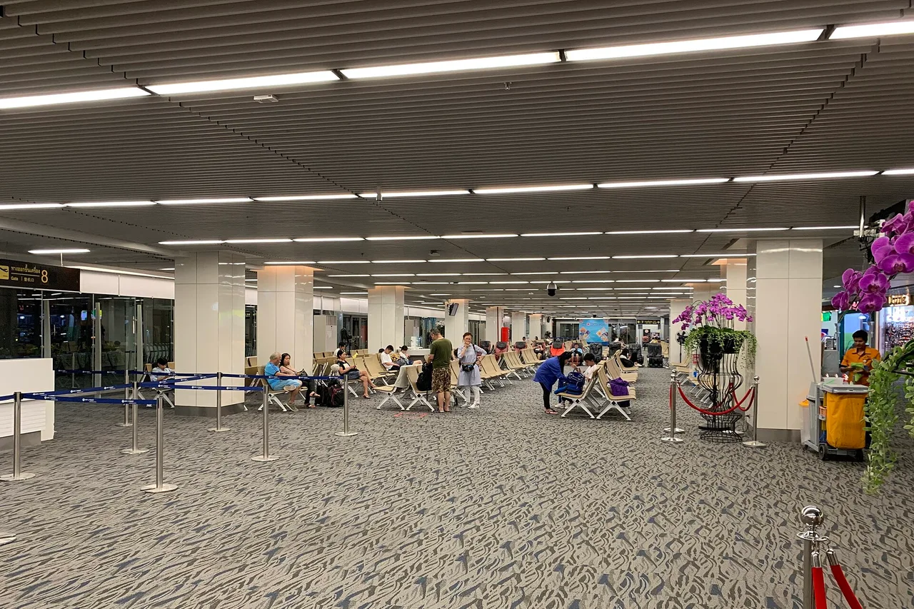 Hell beleuchtete Halle des Inlandsterminals am Flughafen Phuket mit Sitzreihen, Absperrbändern und einer Orchidee