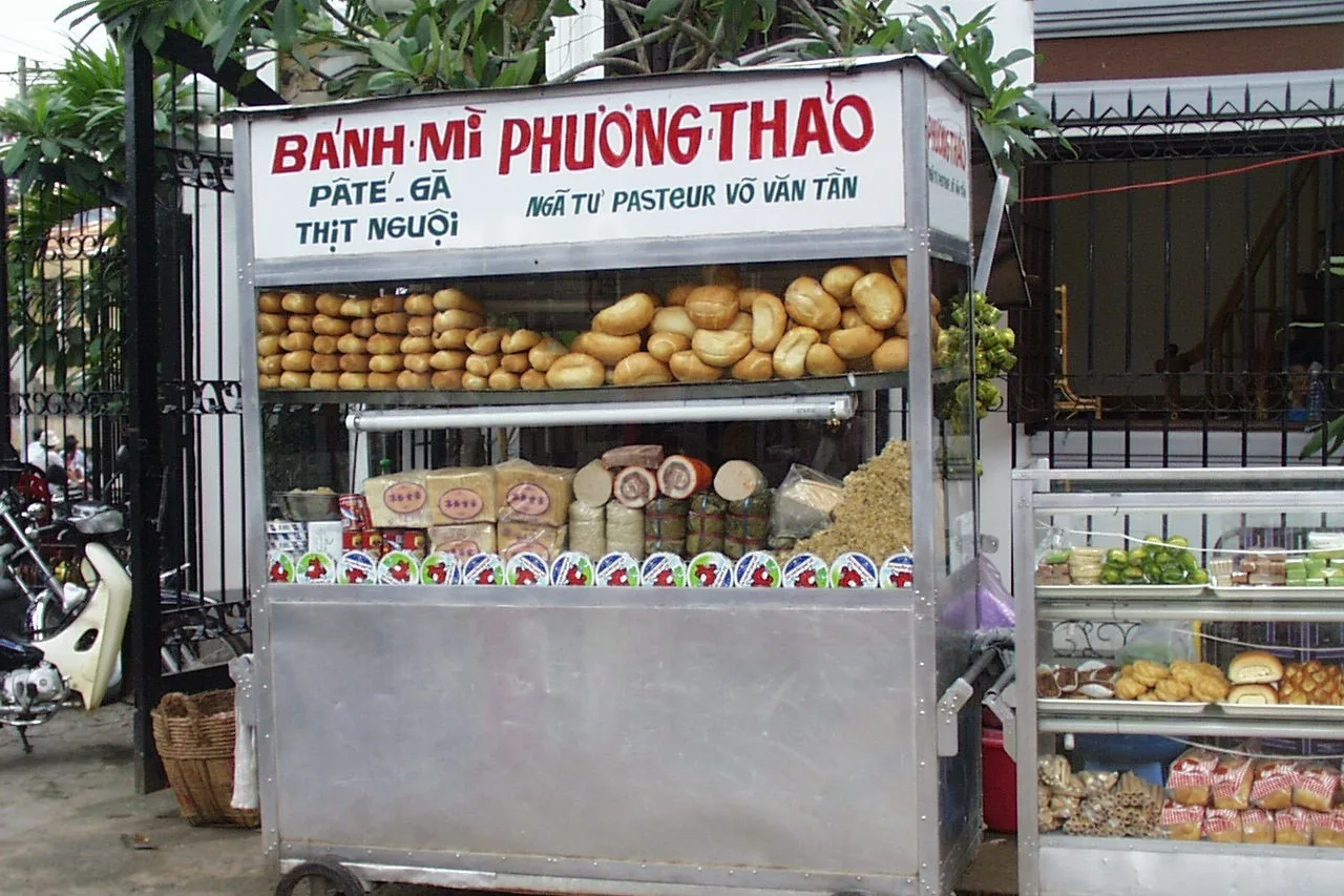 Un carretto di bánh mì con baguette dietro il vetro e l’insegna «Bánh mì Phương Thảo» a bordo strada