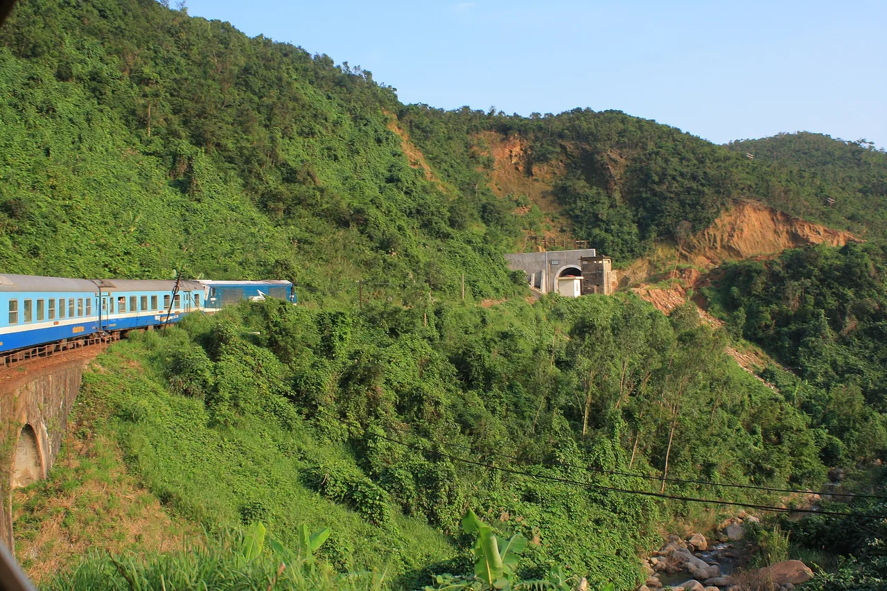 Un treno bianco e blu delle ferrovie vietnamite su un ponte, lungo una collina verde vicino alla costa