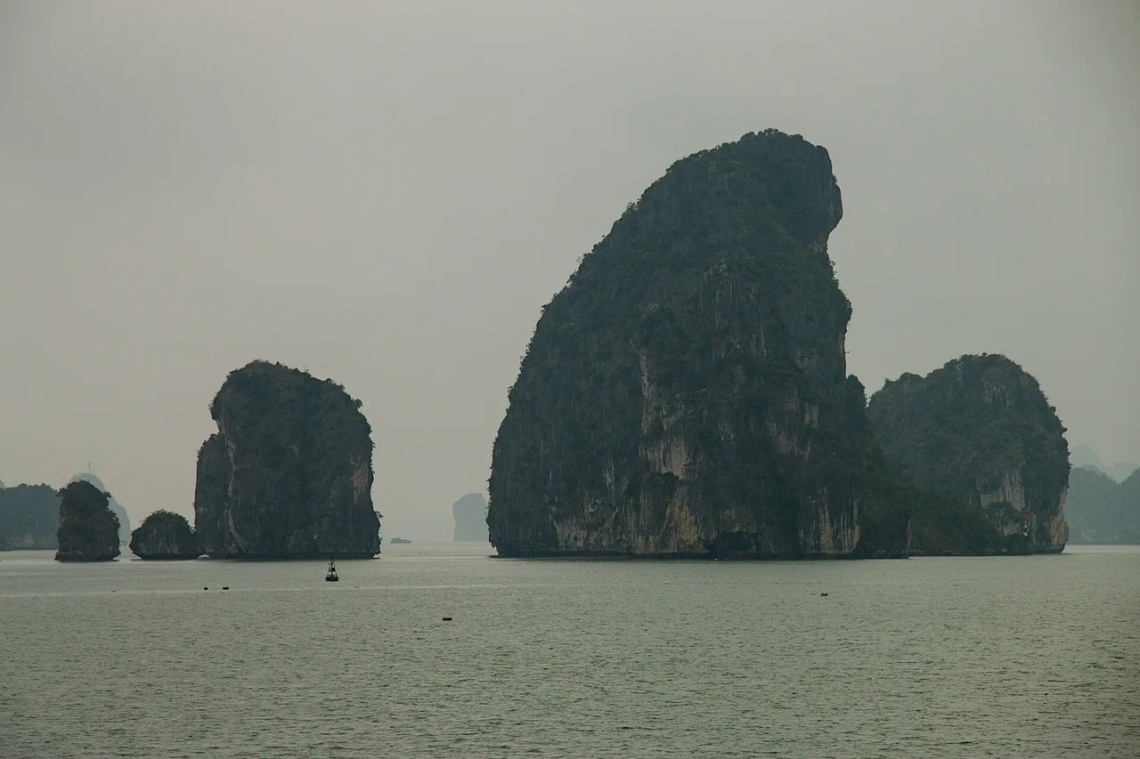 Des rochers calcaires sombres émergent de l'eau grise et calme de la baie de Bái Tử Long sous un ciel couvert