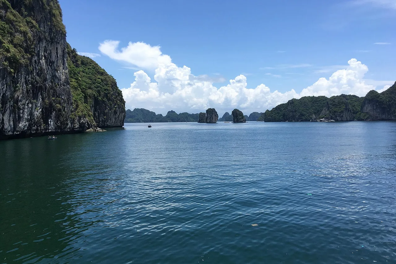 Pitons calcaires verts et eau calme d'un bleu profond dans la baie de Lan Ha, avec d'autres îles de la baie d'Halong à l'horizon sous des nuages blancs