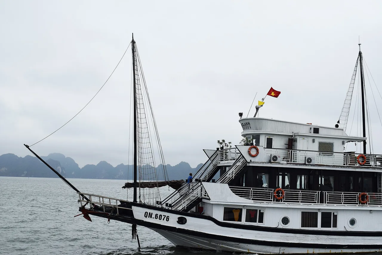 Un bateau de croisière blanc au drapeau vietnamien et une jonque à mât dans la baie d'Halong, des îles calcaires dans la brume en arrière-plan