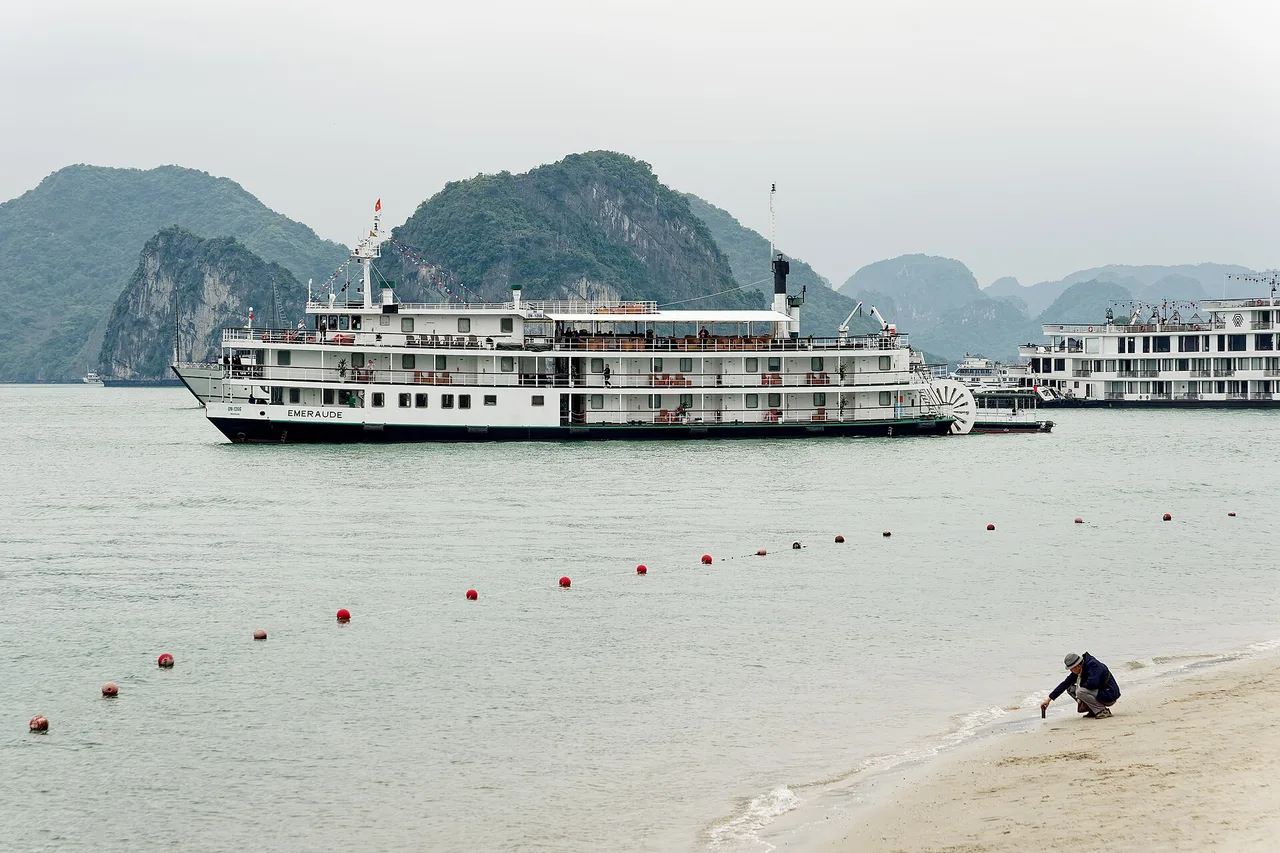 Deux grands bateaux de croisière devant des collines calcaires vertes, vus depuis la plage de l'île de Ti Tốp, une ligne de bouées sur l'eau