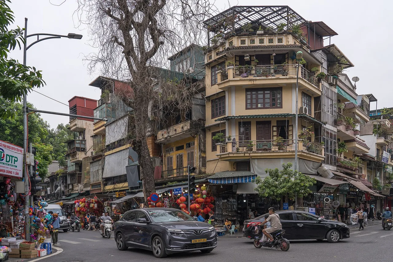 Carrefour du vieux quartier de Hanoï avec des maisons-tubes étroites de plusieurs étages, des arbres, des scooters et des voitures