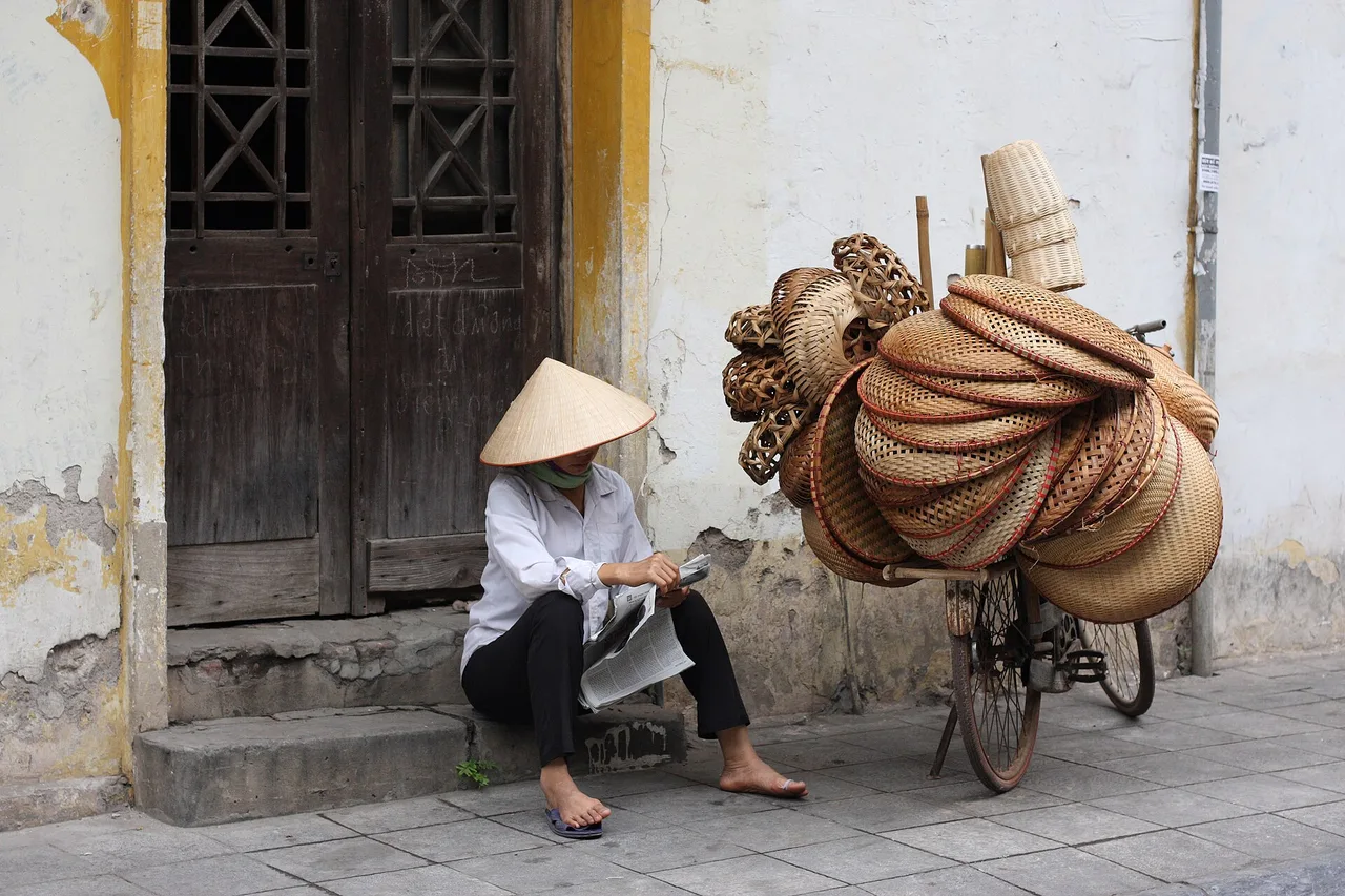Una venditrice con il cappello conico seduta davanti a una vecchia porta di legno nel quartiere vecchio di Hanoi, accanto alla bicicletta carica di cesti intrecciati.