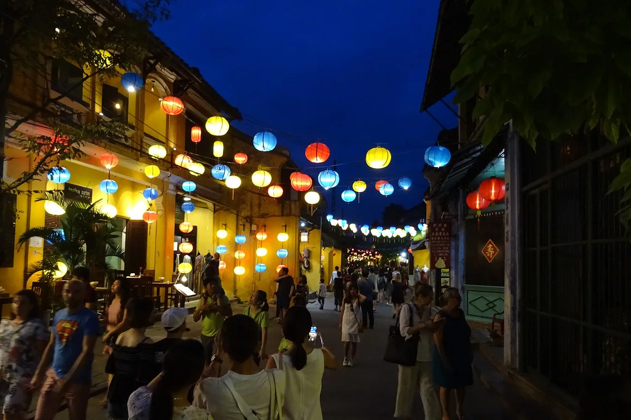 Un vicolo della città vecchia di Hoi An la sera: facciate gialle, lanterne di seta colorate sopra la strada e passanti.