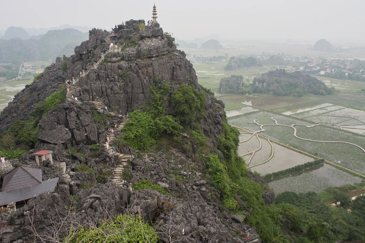 Stone stairway along the ridge of the Hang Mua rock with a small pagoda on the summit, flooded rice fields and limestone hills in the haze below