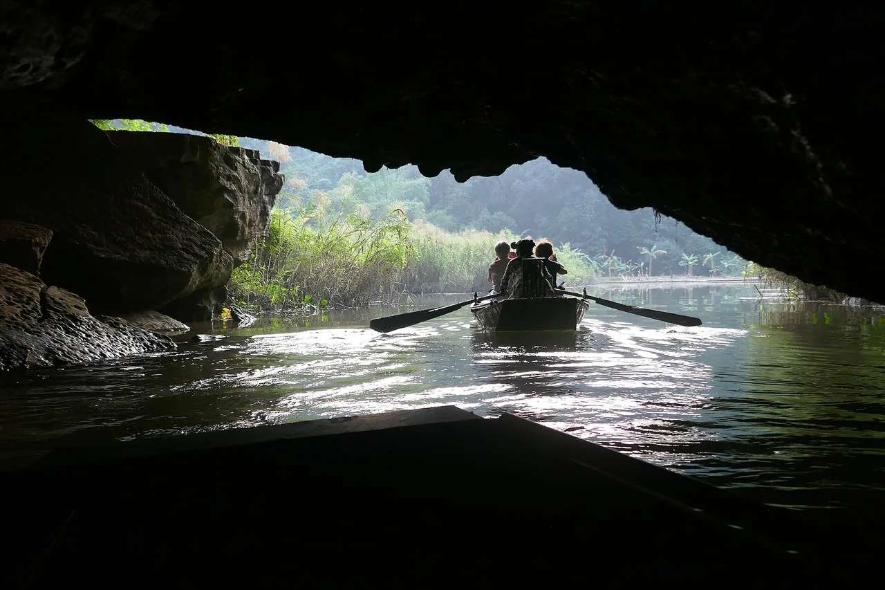 A rowing boat carrying visitors emerges from a dark cave at Tam Coc onto the bright river between limestone cliffs