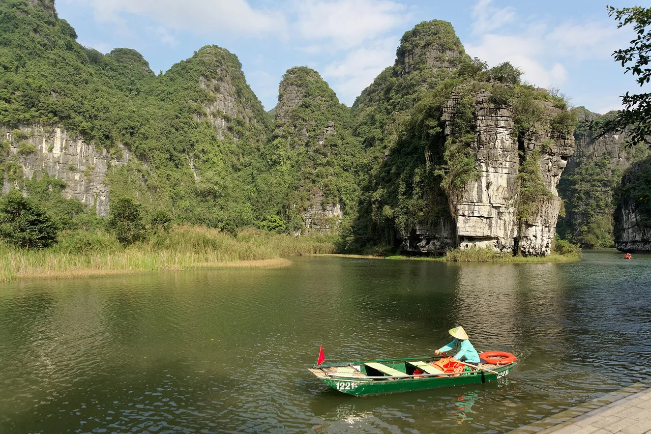 A woman rowing a green metal boat on the calm water of Trang An in front of vegetation-covered limestone cliffs
